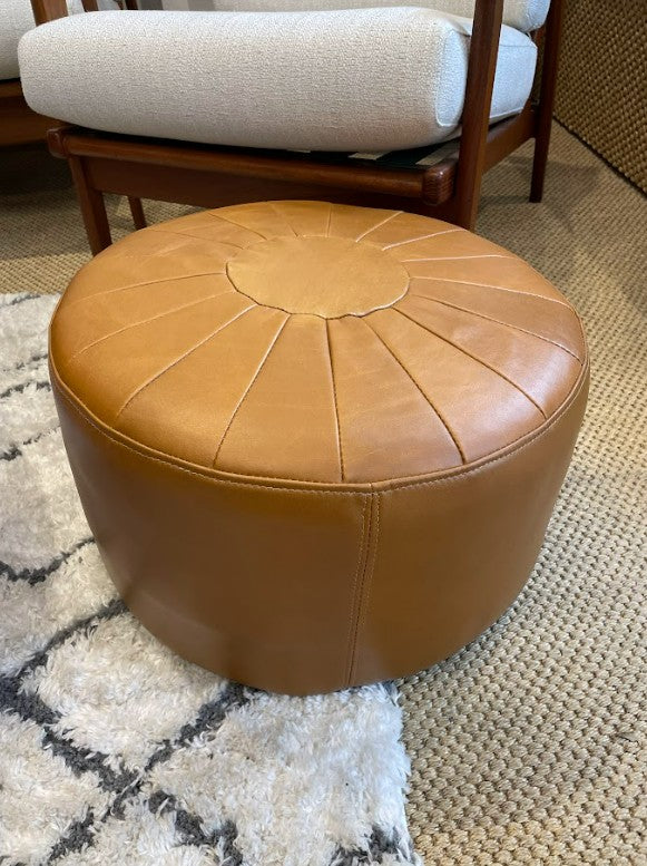 Brown leather ottoman on a textured rug with a chair in the background. Available to purchase online. Ex Parker People™