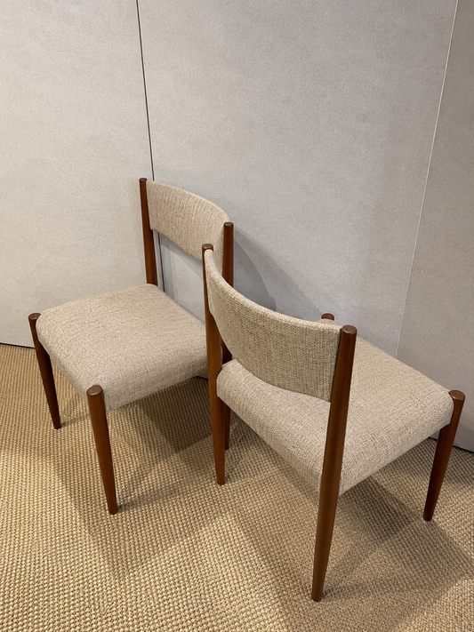 Two mid-century modern dining chairs with beige textured fabric upholstery and dark brown wooden legs, shown on a textured beige carpet against a light gray wall.